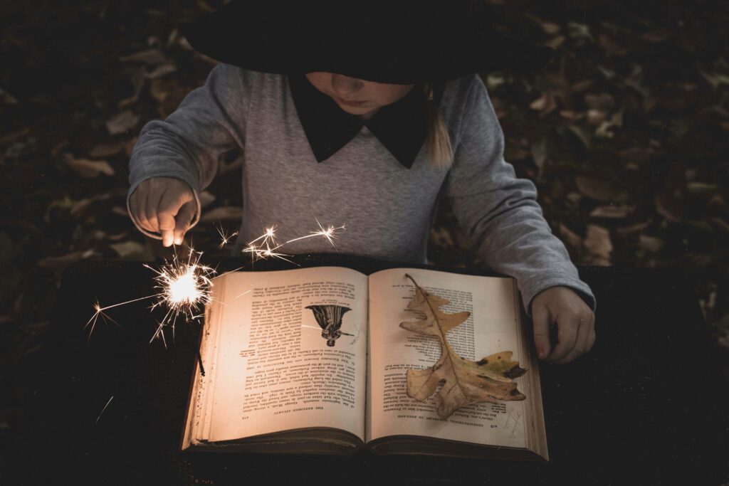 Child wearing a large black hat that hides the eyes. sitting over a book with a burning magic candle in one hand. The book looks old and mysterious, and a wilted oak leaf lies on one page.