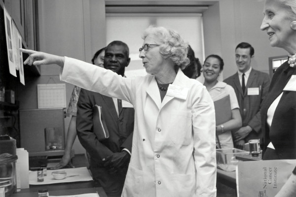Early days of science communication: Black and white photo taken in the 1950s or 60s. An elderly female researcher in a white labcoat points at a paper diagram on the wall, talking to a group of people around her.